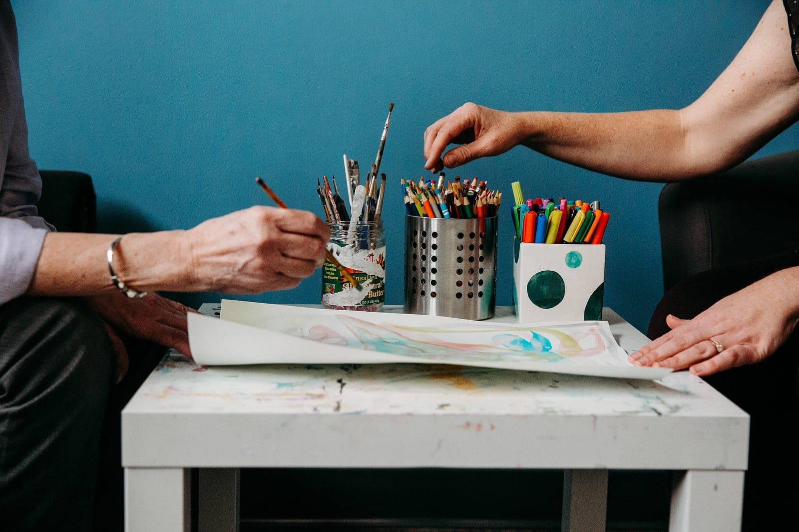 Two people at a paint-splattered studio table filled with jars of paintbrushes, colored pencils, and markers, working on a watercolor painting during a session, a teal wall behind them.